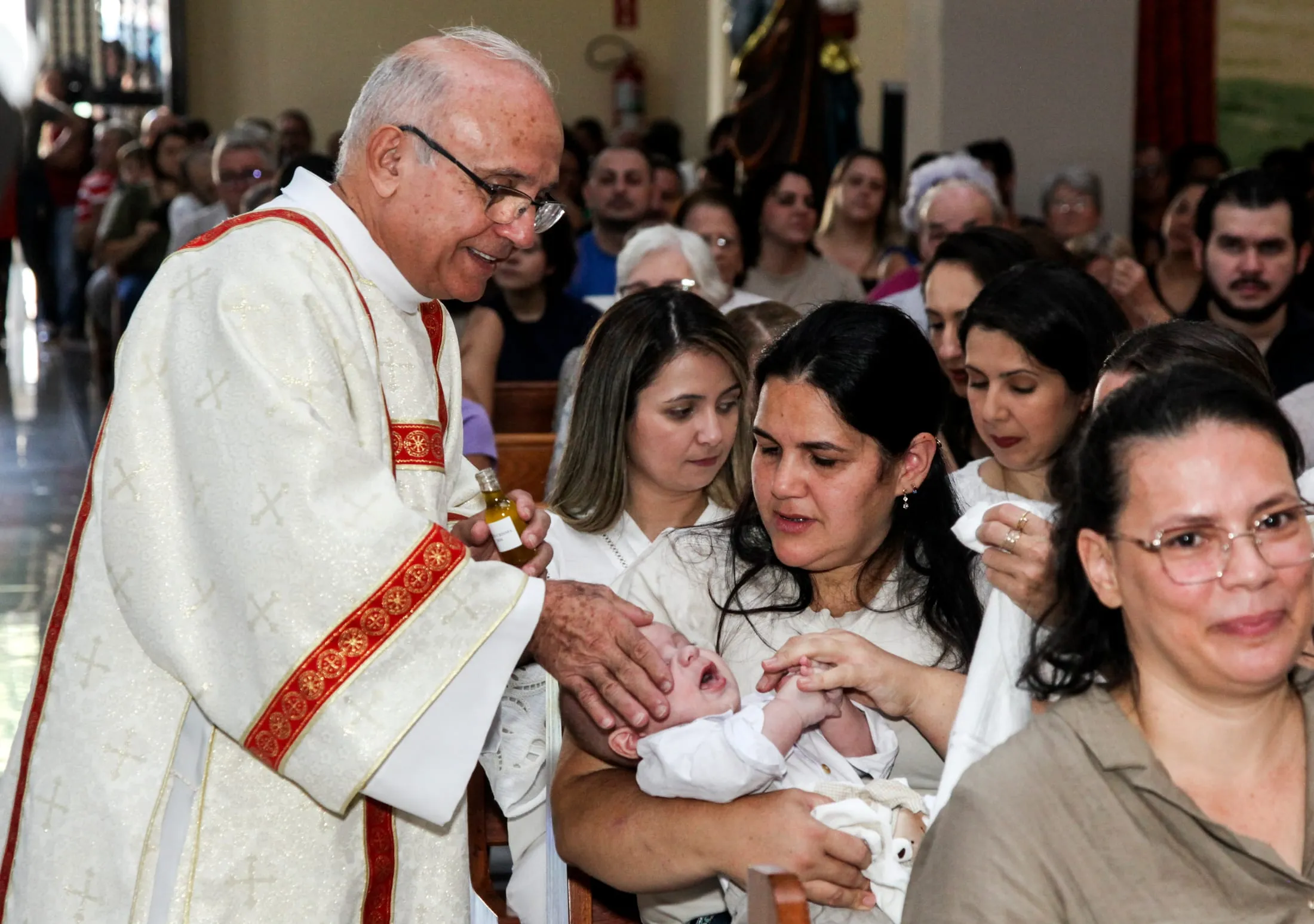 Fotógrafo de batizado em Piracicaba — padre ungindo a testa do bebê entre os fiéis