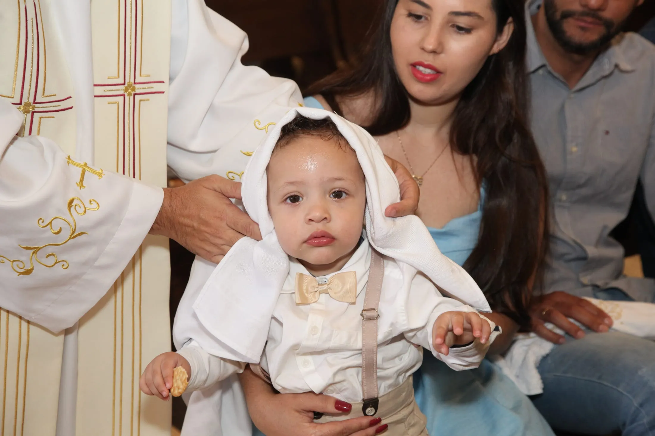 Fotografia de batismo em Piracicaba — padre colocando a veste branca na criança ao lado dos pais